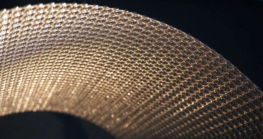 Primo piano di una superficie metallica a maglia dorata, texture a reticolo curvo con riflessi lucidi su sfondo scuro.