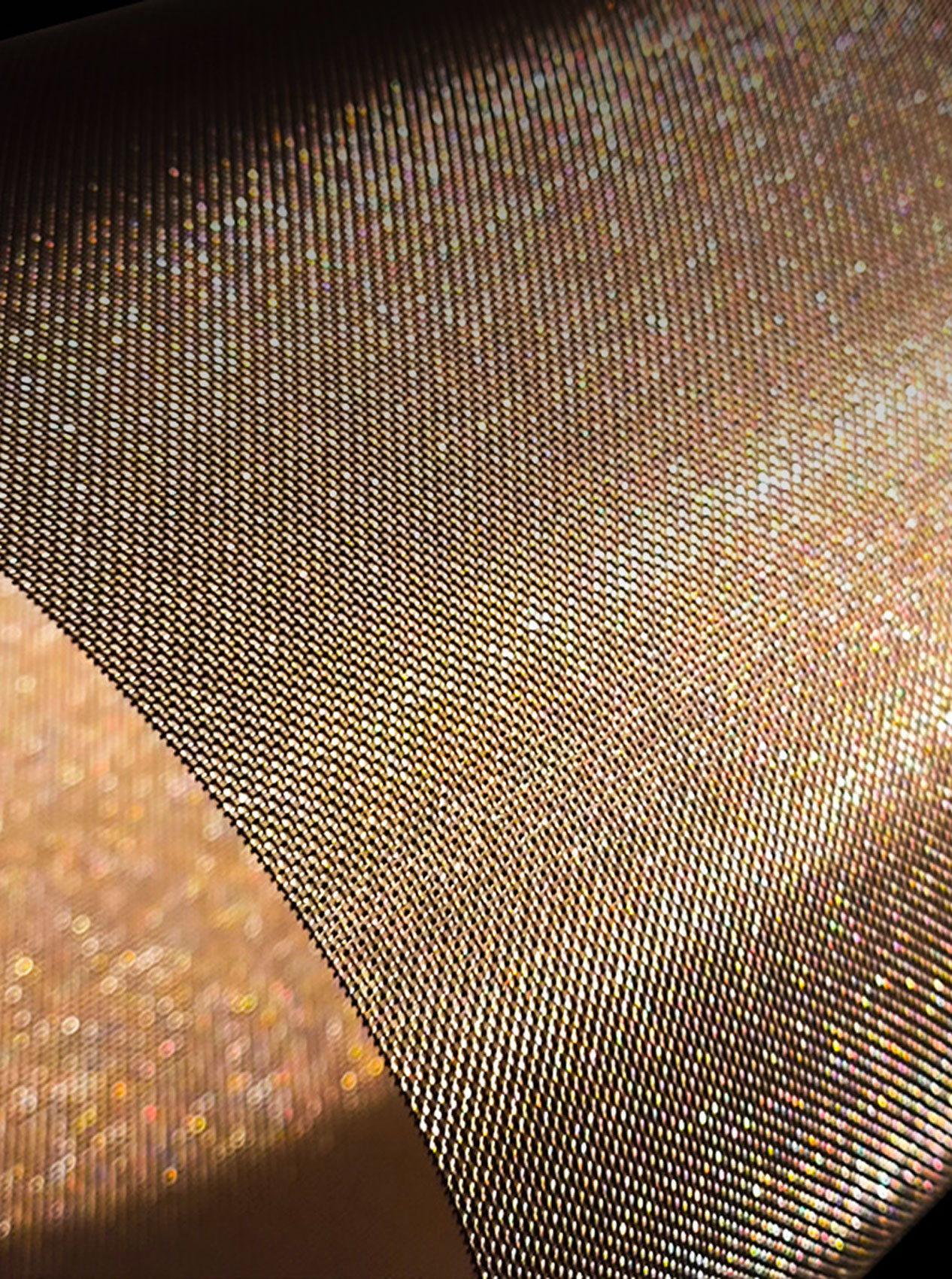 Primo piano di una stoffa dorata lucida con texture a piccoli motivi a righe, riflessi di luce calda e sfumature metalliche.