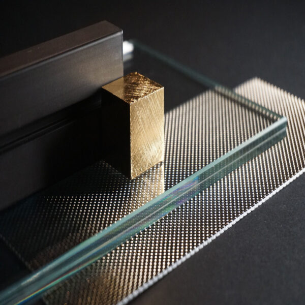 Metallo e vetro: una piccola barretta dorata poggia su una base di vetro e piastrelle metalliche, in un primo piano luminoso.